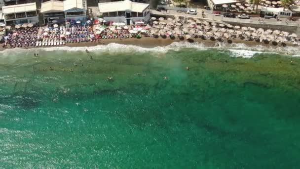 Azureの砂浜の上を飛ぶ空の景色人々はタオルで日光浴をし、緑色の水で遊んでいます. — ストック動画