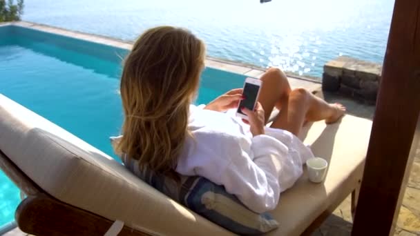 Bela mulher de biquíni sorridente na praia da piscina usando smartphone. Menina mensagens de texto e navegação na internet no telefone móvel, deitado em cadeira de praia na área lateral da piscina do hotel. Banhos de sol e relaxamento no resort — Vídeo de Stock