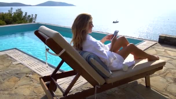 Bela mulher de biquíni sorridente na praia da piscina usando smartphone. Menina mensagens de texto e navegação na internet no telefone móvel, deitado em cadeira de praia na área lateral da piscina do hotel. Banhos de sol e relaxamento no resort — Vídeo de Stock