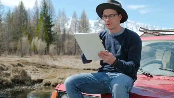 Jeune homme de mode hipster avec tablette d'ordinateur assis à côté de sa voiture pendant le voyage. 20s 4k — Video