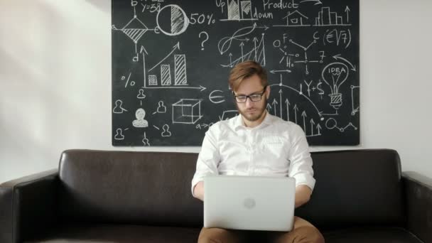 Multitasking. Knappe jonge man een bril en werken met touchpad zittend op de Bank in office 20s 4 k — Stockvideo