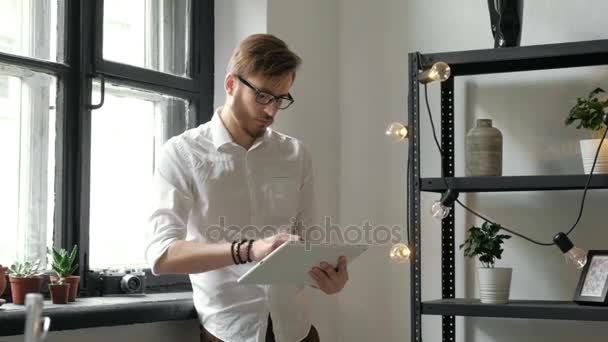 Trabajando en una nueva tableta. Vista de ángulo bajo del joven confiado que trabaja en la tableta digital mientras está parado delante de la ventana grande en la oficina o la cafetería 20s 4k . — Vídeos de Stock