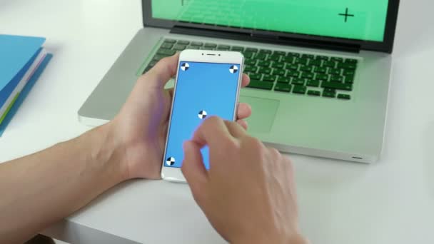 Ділова людина використовує смартфон, перевіряючи інформацію на мобільному телефоні з зеленим екраном на коричневому фоні з ноутбуком. Руки зверху. Тло офісних столів. 20s 4k . — стокове відео