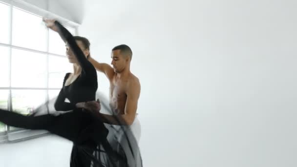 Een balletdanser in wit en een ballerina in zwarte dans in een witte loft naast een groot raam, close-up 20s 4 k. — Stockvideo