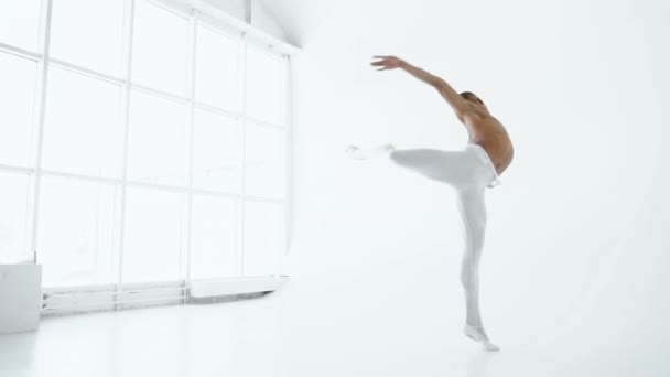 A ballet dancer with a bare torso and white white tights dancing against the backdrop of a large window in a white room — Stock Video