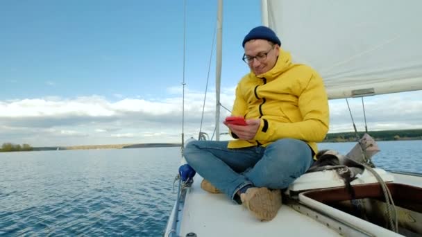 Junger Mann sitzt auf Segelboot und surft Smartphone auf Hintergrund des Meeres — Stockvideo