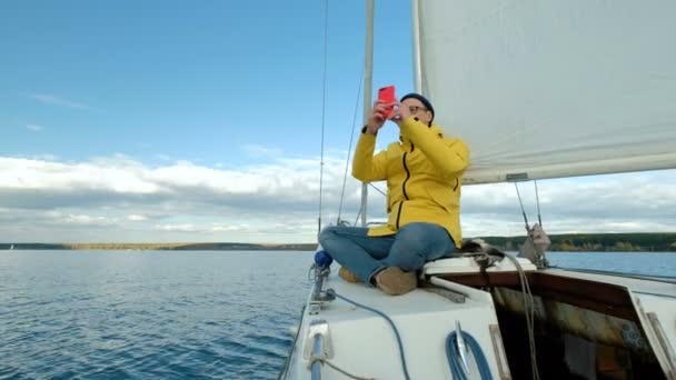 Joven sentado en velero y navegando por teléfono inteligente en el fondo del mar . — Vídeos de Stock