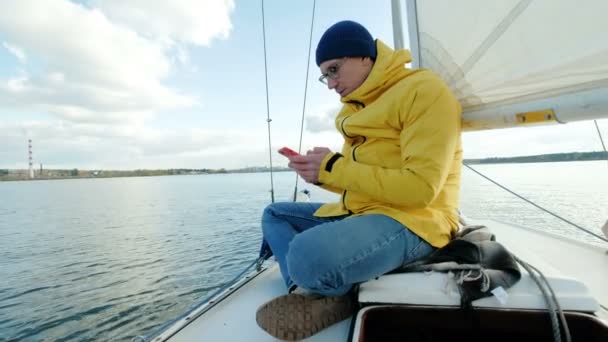 坐在帆船上的年轻人, 在海的背景下浏览智能手机. — 图库视频影像