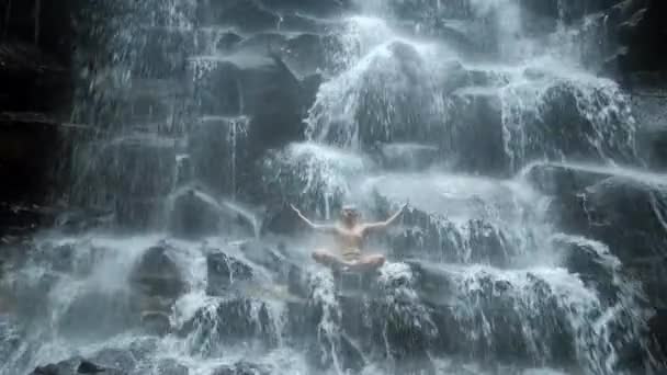 Человек, сидящий в позе йоги на фоне водопада — стоковое видео