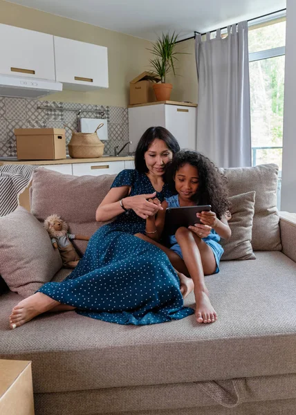 Улыбающаяся мать и ее милая маленькая дочь смотрят что-то на цифровом планшете, сидя вместе на диване в своей гостиной — стоковое фото