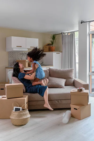 Mamá e hija multirraciales se mudan a un nuevo apartamento. Diviértete saltando y jugando en el sofá . — Foto de Stock