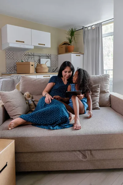Mosolygó anya és az ő aranyos kis lánya nézi valamit a digitális tabletta ülve együtt a nappaliban kanapé — Stock Fotó