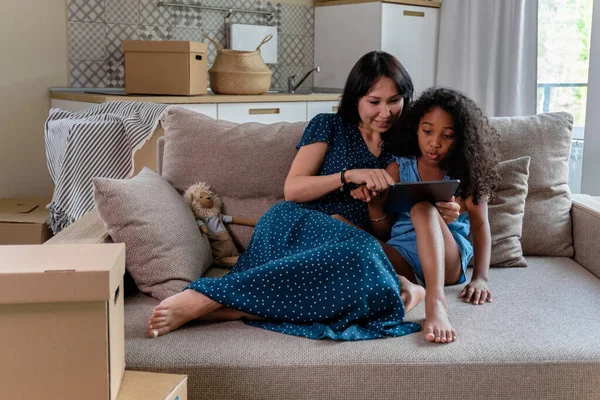 Leende mamma och hennes söta lilla dotter tittar på något på en digital tablett när du sitter tillsammans på deras vardagsrum soffa — Stockfoto