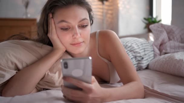 Mujer relajada en casa leyendo un mensaje de texto en su dormitorio luminoso. Mujer en la cama revisando aplicaciones sociales con smartphone — Vídeos de Stock