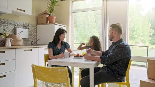 Mooie familie met Kid zittend aan de tafel koffie drinken in nieuw huis rond kartonnen dozen — Stockvideo