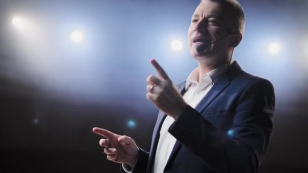 Showman. Middle Age Male entertainer, presenter or actor on stage. Arms to sides, smoke on background of spotlight. Front view portrait of a male public speaker speaking at the microphone, pointing — Stock Video