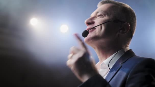 Portret van mannen die spreken via een microfoon in de donkere conferentiezaal. Hij praat tegen microfoons op een persconferentie. Profiel van volwassen mannelijke politicus spreekt hartstochtelijk vanuit tribune — Stockvideo