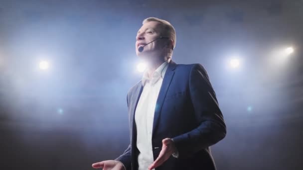 Showman. Middle Age Male entertainer, presenter or actor on stage. Arms to sides, smoke on background of spotlight. Front view portrait of a male public speaker speaking at the microphone, pointing — Stock Video