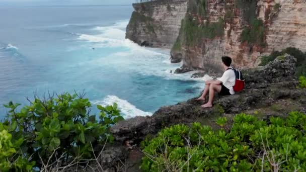 El tipo se sienta al borde del acantilado. Un joven que llega al borde del acantilado y se sienta. Hombre sentado y mirando el horizonte. El chico llega al borde del peñasco y se sienta en la roca. Solitario sentado sobre el acantilado — Vídeos de Stock