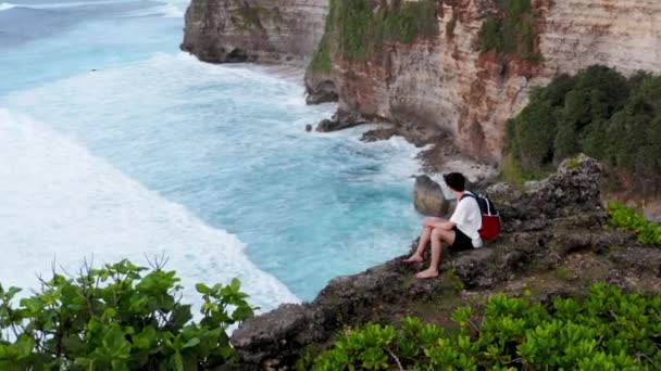 El tipo se sienta al borde del acantilado. Un joven que llega al borde del acantilado y se sienta. Hombre sentado y mirando el horizonte. El chico llega al borde del peñasco y se sienta en la roca. Solitario sentado sobre el acantilado — Vídeo de stock