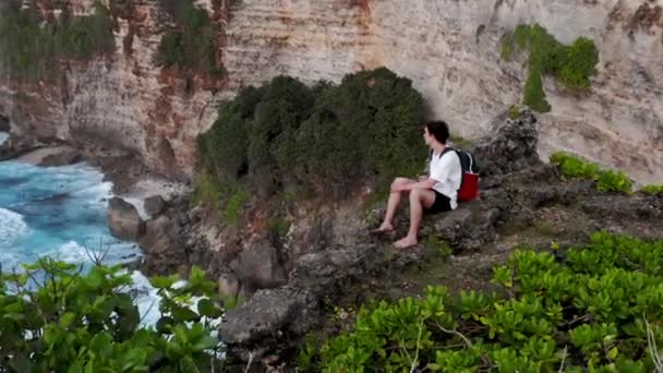 那家伙坐在悬崖边上。小男孩来到悬崖边，坐下。人坐着看着地平线。男孩来到峭壁的边缘，坐在岩石上。孤独的家伙坐在悬崖上 — 图库视频影像