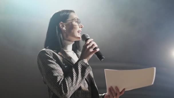 La entrenadora financiera habla emocionalmente desde el escenario con los espectadores en el foro. Demasiadas personas anónimas trabajadores y estudiantes se sientan en un gran auditorio y observan los antecedentes del taller — Vídeos de Stock