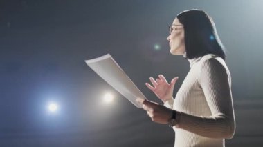 Kadın finans koçu, forumda seyircilerle sahnede konuşurken duygusal jestler yapıyor. Birçok isimsiz insan ve öğrenci büyük bir salonda oturup atölye çalışmalarını izliyor.