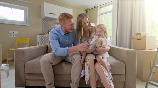 Glad sød lille pige køre udforske nyt hus, mens forældre omfavne på sofaen, familie med aktivt barn datter lege på bevægende dag koncept – Stock-video