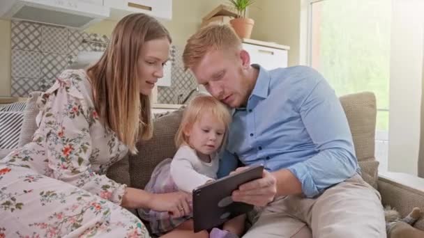 Alegre familia caucásica con la niña sentada en el sofá, riendo mientras ve algo muy divertido en el dispositivo de la tableta. En casa. Mirando Positivo a la reubicación o desembalaje del paquete de cartón . — Vídeos de Stock