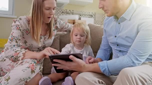 Vrolijke blanke familie met een klein meisje op de bank, lachend terwijl ze iets heel grappigs zag op het tablet apparaat. Thuis. Positief kijken naar het verplaatsen of uitpakken van het kartonnen pakje. — Stockvideo
