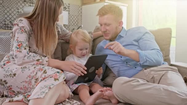 Famiglia caucasica allegra con bambina seduta sul divano, che ride guardando qualcosa di molto divertente sul dispositivo tablet. A casa. Uno sguardo positivo alla ricollocazione o allo spacchettamento del pacchetto di cartone . — Video Stock