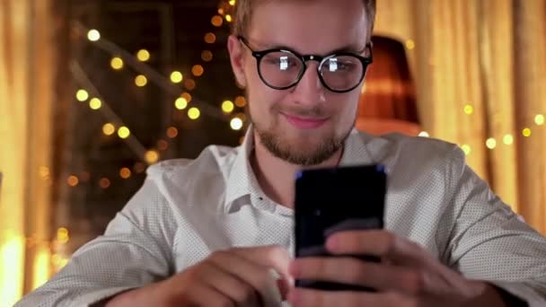 Knappe man met een baard die een smartphone gebruikt en glimlacht terwijl hij bier drinkt in de pub — Stockvideo