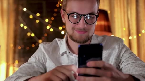 Bonito homem barbudo usando um smartphone e sorrindo enquanto bebe cerveja no pub — Vídeo de Stock