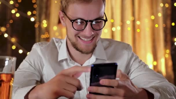 Bonito homem barbudo usando um smartphone e sorrindo enquanto bebe cerveja no pub — Vídeo de Stock
