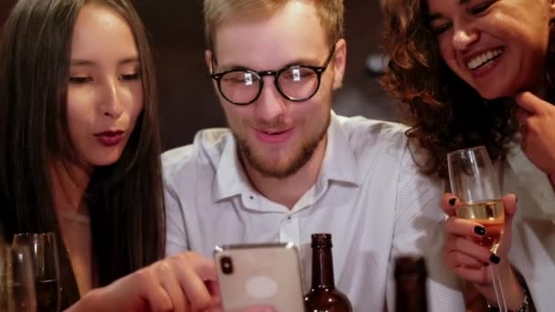 Retrato de grupo de estudiantes hipster felices viendo video, foto en el teléfono inteligente juntos mientras están sentados en la mesa de madera en el bar moderno, pub . — Vídeo de stock