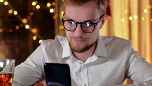 Hombre barbudo guapo usando un teléfono inteligente y sonriendo mientras bebe cerveza en el pub — Vídeos de Stock