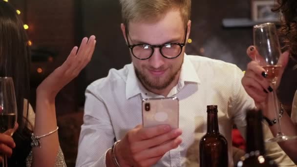 Amigos en un pub celebran y se divierten, un tipo está mirando un teléfono inteligente, y no hablar con otras personas. El problema de la sociedad moderna . — Vídeo de stock