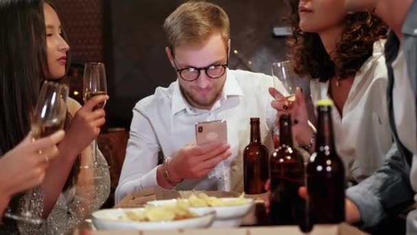 Amigos em um pub celebram e se divertem, um cara está olhando para um smartphone e não falando com outras pessoas. O problema da sociedade moderna . — Vídeo de Stock