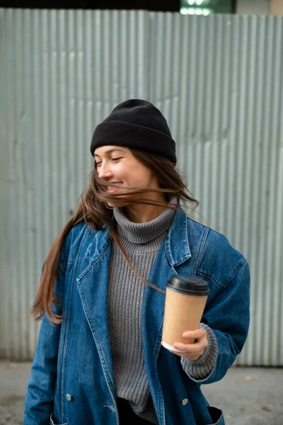 Серйозно виглядає красива молода жінка, позує на камеру на вулиці в місті, одягнена в стильне вбрання, під час виходу на вулицю, тримаючи паперову чашку з кавою — стокове фото