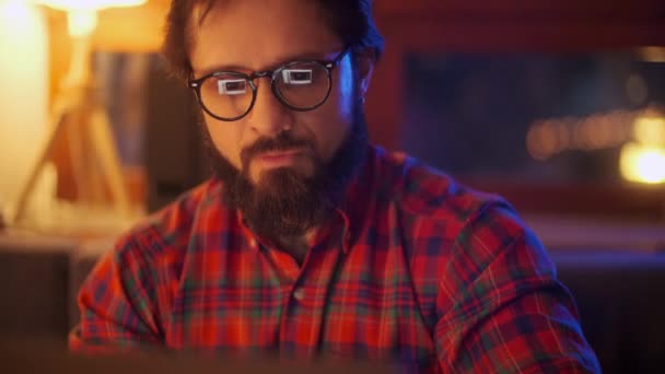 Close Up Retrato de um jovem empresário barbudo vestindo óculos sentado em seu escritório trabalhando tarde à noite olhando para monitor em um computador. Tela do computador reflete em seus óculos . — Vídeo de Stock