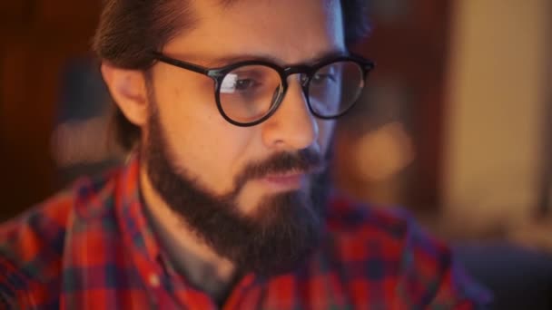 Närbild Porträtt av en skäggig ung affärsman bär glasögon sitter i sitt kontor arbetar sent på natten tittar på bildskärm på en dator. Datorskärmen reflekterar i hans glasögon. — Stockvideo