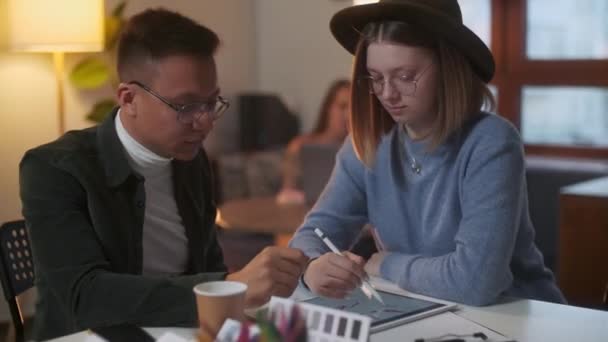 Młodzi stylowi projektanci omawiają projekt na tablecie rysując długopisem, tłem Kobieta i mężczyzna z laptopem. — Wideo stockowe