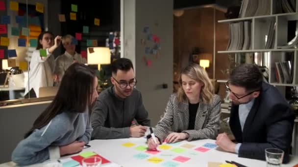 Les designers de l'équipe créative travaillent sur un nouveau projet, le partage d'idées, l'écriture d'informations sur des autocollants colorés. Mur de bureau en verre avec des mémos lumineux est en arrière-plan . — Video