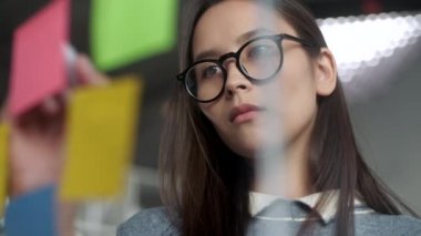 Genç İş Kadını Beyin Fırtınası. Cam duvara yapıştırılmış yapışkan notlara fikir yazan Asyalı kadın. İş Başarı Konsepti