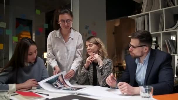 Команда креативных дизайнеров смотрит на журнал Project, говорит о них, сидит вместе за столом. Современный неформальный офис. Дружественная рабочая атмосфера — стоковое видео