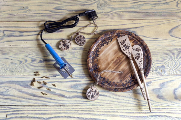 A set of tools for burning wood with nozzles and examples of wooden objects with drawings on a wooden table close-up