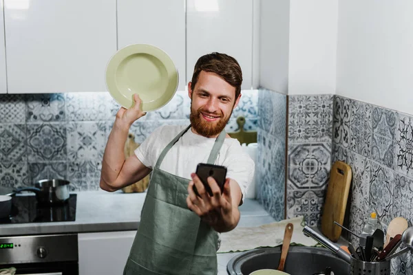 A young bearded man holds a washed plate and takes a selfie on the phone.