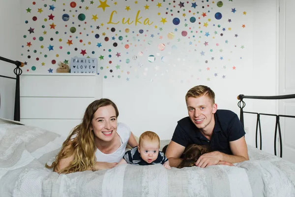 Young mom and dad are lying with the baby on the bed, smiling and looking at the camera.