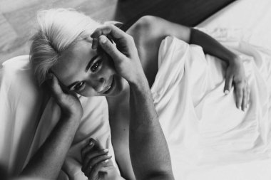 Genç bir adam ve bir kadın yatakta sarılıyor. Bir erkek yatakta bir kadına bakar..