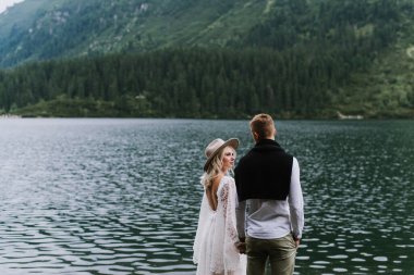 Boho elbisesi içinde güzel bir gelin, bir şapka ve damadın kucaklaşması ve dağlarda bir gölün kenarında durması. Gelin ve damat dağlarda kucaklaşır.